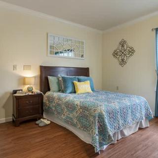 A cozy bedroom with a double bed featuring a colorful quilt, a wooden nightstand with a lamp, and decorative wall art.