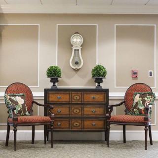 A traditional interior setting featuring two red upholstered chairs flanking a wooden chest of drawers, adorned with decorative plants and a wall-mounted art piece above.