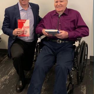 Two people are posing together indoors, one holding a book and the other seated in a wheelchair.