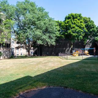 A serene outdoor space featuring lush trees and a grassy area, bordered by buildings under a clear blue sky.