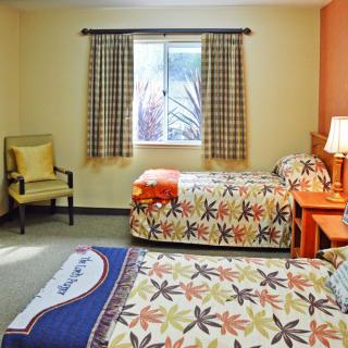 A cozy bedroom with two twin beds featuring colorful floral bedspreads, a window with checkered curtains, a lamp on the bedside table, and an upholstered chair in the corner.