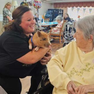 A caregiver shares a joyful moment with a senior resident, holding a small brown animal in a nursing home setting.
