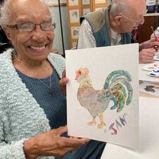 An elderly woman smiles while showing her painting of a rooster in an art class.