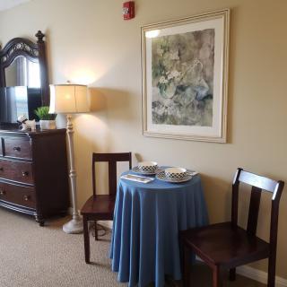 A cozy interior space featuring a small table with a blue tablecloth, two wooden chairs, a floor lamp, and framed artwork on the wall.