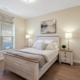 A modern bedroom with neutral tones, featuring a bed with gray and white linens, two nightstands with lamps, and a piece of artwork on the wall.