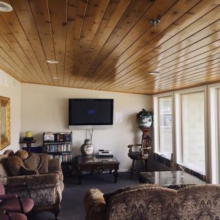 A spacious living room with a wooden ceiling, featuring a television, comfortable sofas, and large windows.