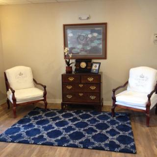 A cozy waiting room with two upholstered chairs, a decorative rug, and a chest of drawers under a painting.