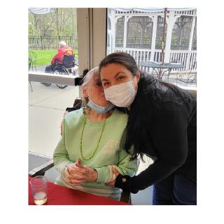 A person wearing a mask leans in close to another person seated in a wheelchair, both inside a room.