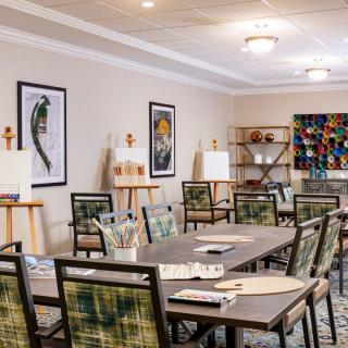 A well-lit art room with tables, chairs, easels, and vibrant paintings on the walls.
