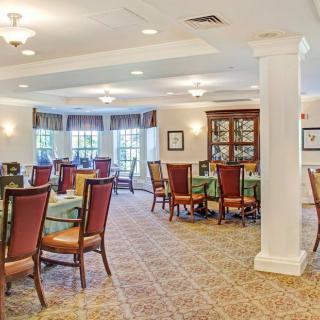 A well-lit dining room with multiple tables and chairs, featuring large windows and a carpeted floor.