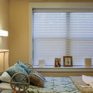 A cozy bedroom featuring a neatly made bed, decorative pillows, and a window adorned with blinds, complemented by personal photographs on display.