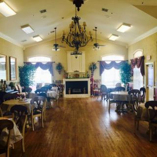 A cozy dining area with elegantly arranged tables and a chandelier, featuring a fireplace and decorative plants.