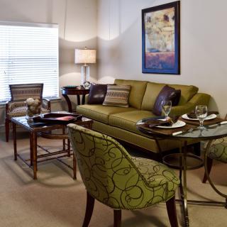 A cozy and elegantly furnished living room with a green sofa, patterned chairs, glass tables, and decorative artwork on the wall.