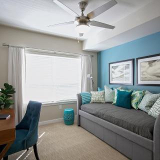 A cozy bedroom with a daybed featuring blue accents and decorative pillows, complemented by a desk and ceiling fan.