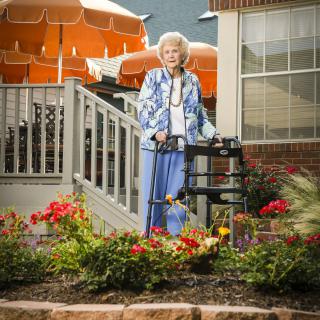 An elderly woman standing with a walker in a garden outside a residential area.