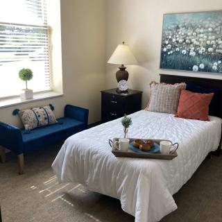 A cozy bedroom featuring a single bed with white linens, a blue bench near the window, and decorative pillows. A lamp and artwork add to the room's ambiance.