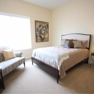 A cozy bedroom with neutral tones, featuring a bed, chair, and wall artwork.
