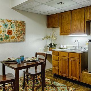 A small kitchen and dining area featuring wooden cabinets, a table with chairs, decorative wall art, and a cozy atmosphere.