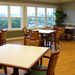 A bright dining room with several tables and chairs, featuring large windows and a wooden floor.