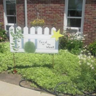 A garden with a 'Yard of the Week' sign next to a brick building.