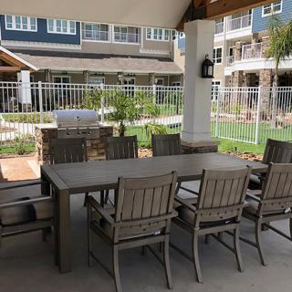 An outdoor patio area featuring a large dining table with chairs, situated near a garden and building.