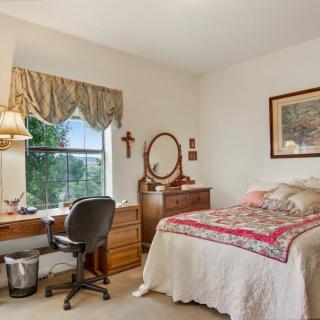 A cozy bedroom with a neatly made bed, a desk by the window, and a dresser topped with a mirror.