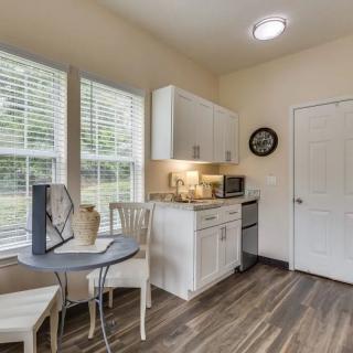 A compact kitchen area with a small dining table and chairs, featuring white cabinets, a microwave, and large windows.