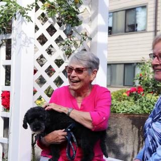 Two smiling women holding a small black dog in a garden setting.