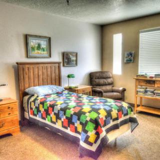 A cozy bedroom featuring a quilted bedspread, a comfortable chair, and decorative artwork.