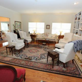 A cozy living room with traditional furniture, featuring armchairs and a sofa around a coffee table on a patterned rug, complemented by hardwood flooring and well-organized bookshelves.