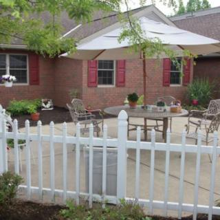 A cozy patio with a table and chairs, shaded by an umbrella, enclosed by a white picket fence in front of a brick house.