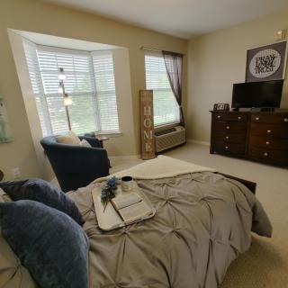 A cozy bedroom with a neatly made bed, featuring blue accent pillows and a tray. The room includes a TV on a dresser and a comfortable chair by the window.