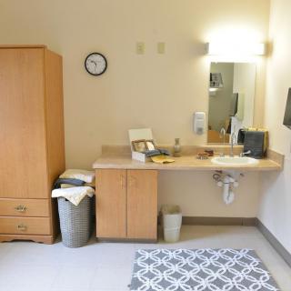 A simple room interior with a wooden dresser, a sink with a mirror above it, a clock on the wall, and a patterned rug on the floor.