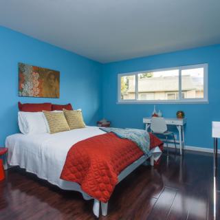 A cozy bedroom with blue walls, featuring a bed with colorful pillows and a desk by the window.