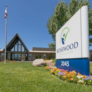 A building with a sign in front displaying 'ROSEWOOD' and colorful flowerbeds under a clear blue sky.