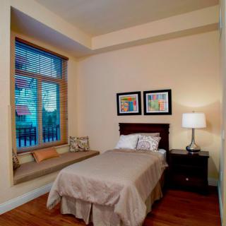 A cozy bedroom with a single bed and window seat, featuring a nightstand with a lamp and colorful artwork on the wall.