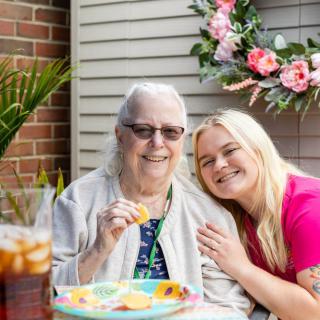 An elderly woman and a younger woman smiling together outdoors, enjoying a snack by a floral arrangement.