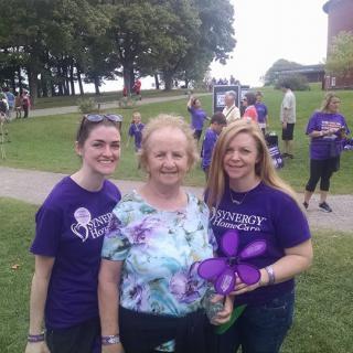 A group of three people at an outdoor event, two wearing purple shirts and holding a flower prop.