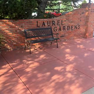 A bench in front of a brick wall labeled 'Laurel Gardens' with sunlight creating shadows on the ground.