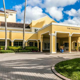 A yellow hotel building with a paved driveway, surrounded by palm trees under a cloudy sky.