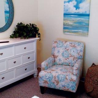 A cozy living room corner featuring a floral armchair, white dresser, mirror, and an ocean-themed painting.