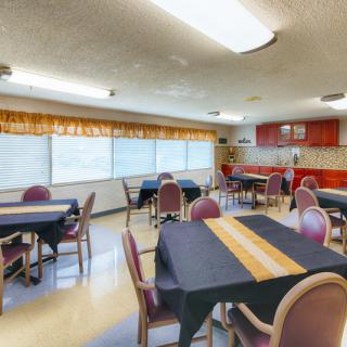 A dining area with multiple tables and chairs, featuring large windows and a kitchen space in the background.