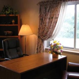 A cozy office setup with a desk, chair, lamp, and window, adorned with curtains and flowers.