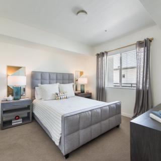 A modern bedroom with a sleek design, featuring a gray padded bed, side tables with lamps, and a large window allowing natural light.