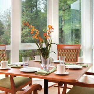 A dining table set with plates, cups, and a flower vase, surrounded by wooden chairs in a bright room with large windows.