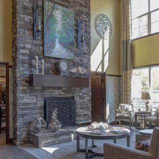 A cozy living room with a stone fireplace and high ceilings, featuring artwork and ample natural light.