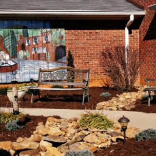 A serene garden scene with a bench in front of a brick wall adorned with a colorful mural and surrounded by neatly arranged rocks and plants.