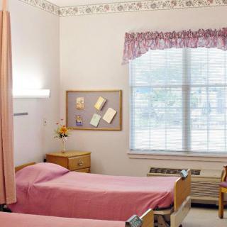 A clean and simple hospital room featuring two beds with pink linens, a window with floral curtains, and a bulletin board on the wall.