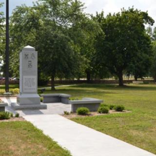 A monument stands in a park setting, surrounded by trees and a neatly trimmed lawn.