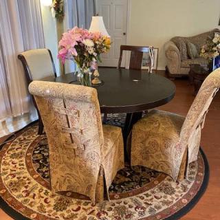 A cozy dining room featuring a round table with upholstered chairs, floral centerpiece, and a decorative carpet.
